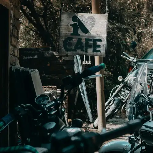 Rustic wooden “I Love Cafe” sign hanging outdoors with motorbikes parked nearby.