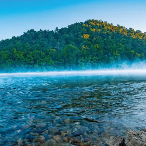 Calm blue water of the Kosi River with mist and a forested hill in the background near Ramnagar.
